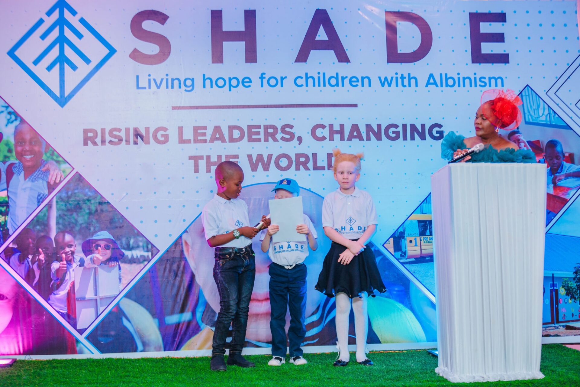 children standing on a stage wearing “SHADE” t-shirts, some holding microphones, participating in a performance or presentation.